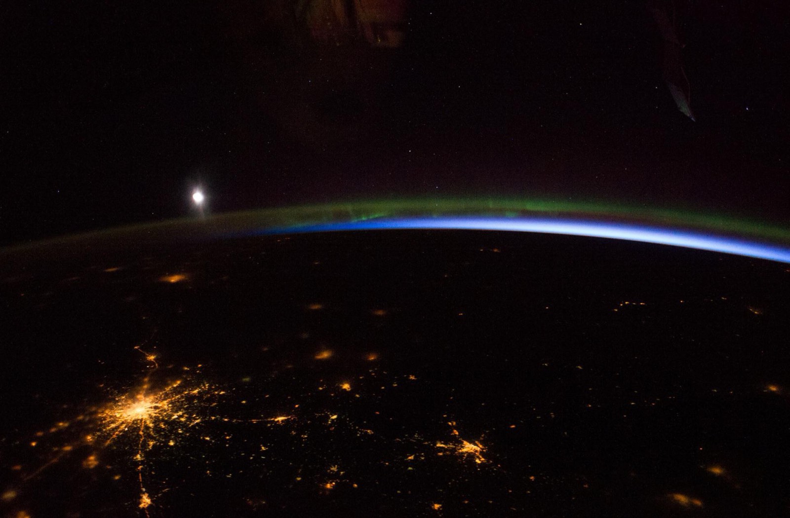 Moscow at night from orbit: yellow radial city lights, a green aurora line above the Earth's limb, and the moon as a white disc